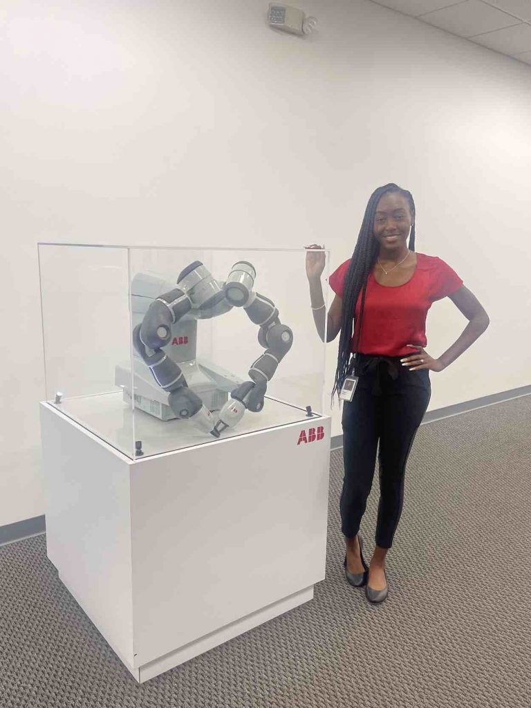 Tishira stands in a red shirt and black pants next to display at global technology company, ABB.