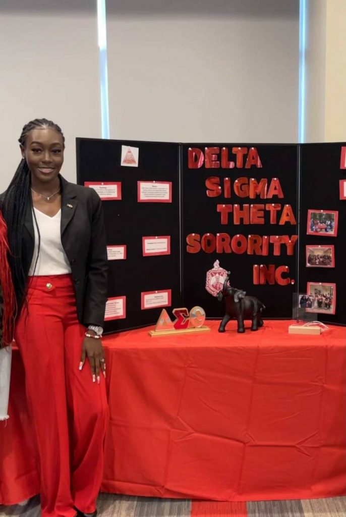 Tishira wears red pants and a black blazer in front of a poster for a Delta Sigma Thera Sorority presentation.