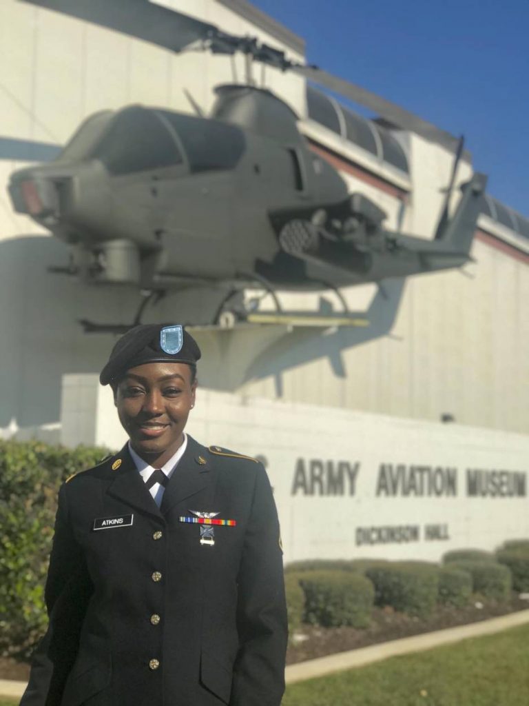Tishira stands in a military uniform in front of the U.S. Army Aviation Museum with a helicopter on display.