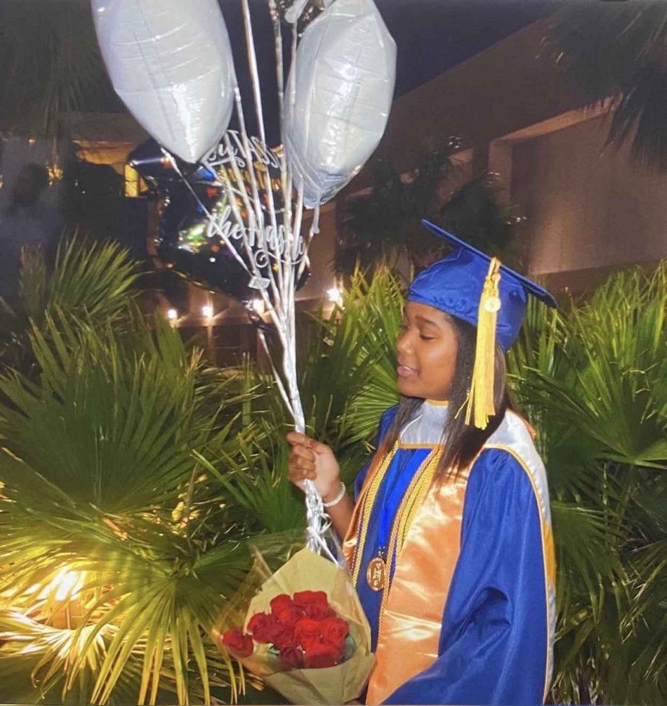 Cambria stands in a blue graduation cap and gown with red roses and white balloons.