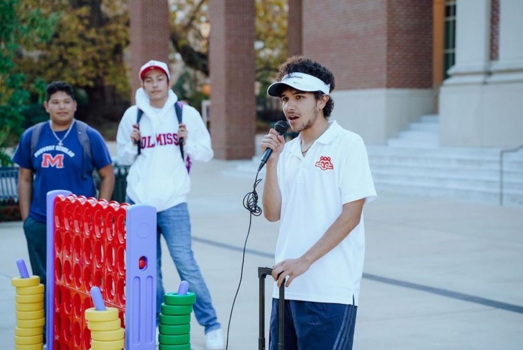 Andy Flores stands in a white shirt and visor and speaks to a crowd with a microphone in front of the Ole Miss Student Union.