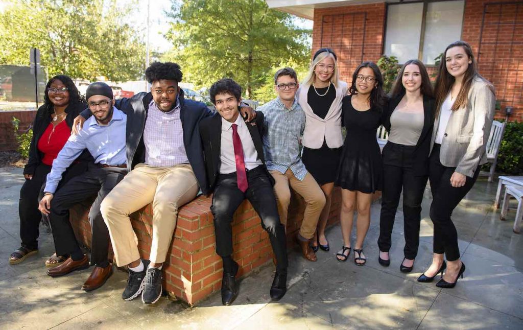 Andy Flores wears a black suit and a red tie as he sits in front of a brick buillding with other members of the First Generation Student Network.