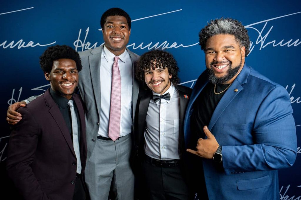 Andy Flores, in a black suit with a bow tie, poses in front of a blue screen with three other gentlemen in suits.