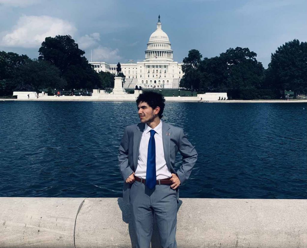 Andy Flores stands in a gray suit and blue tie in Washington, D.C. in front of the water feature with the the U.S. Capitol in the background.