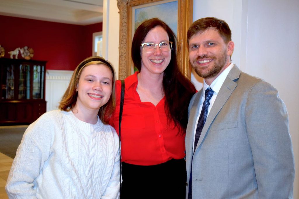 Jamie poses in a suit and tie with his wife and daughter. His daugther is on the left, his wife is in the middle and Jamie is on the right. 