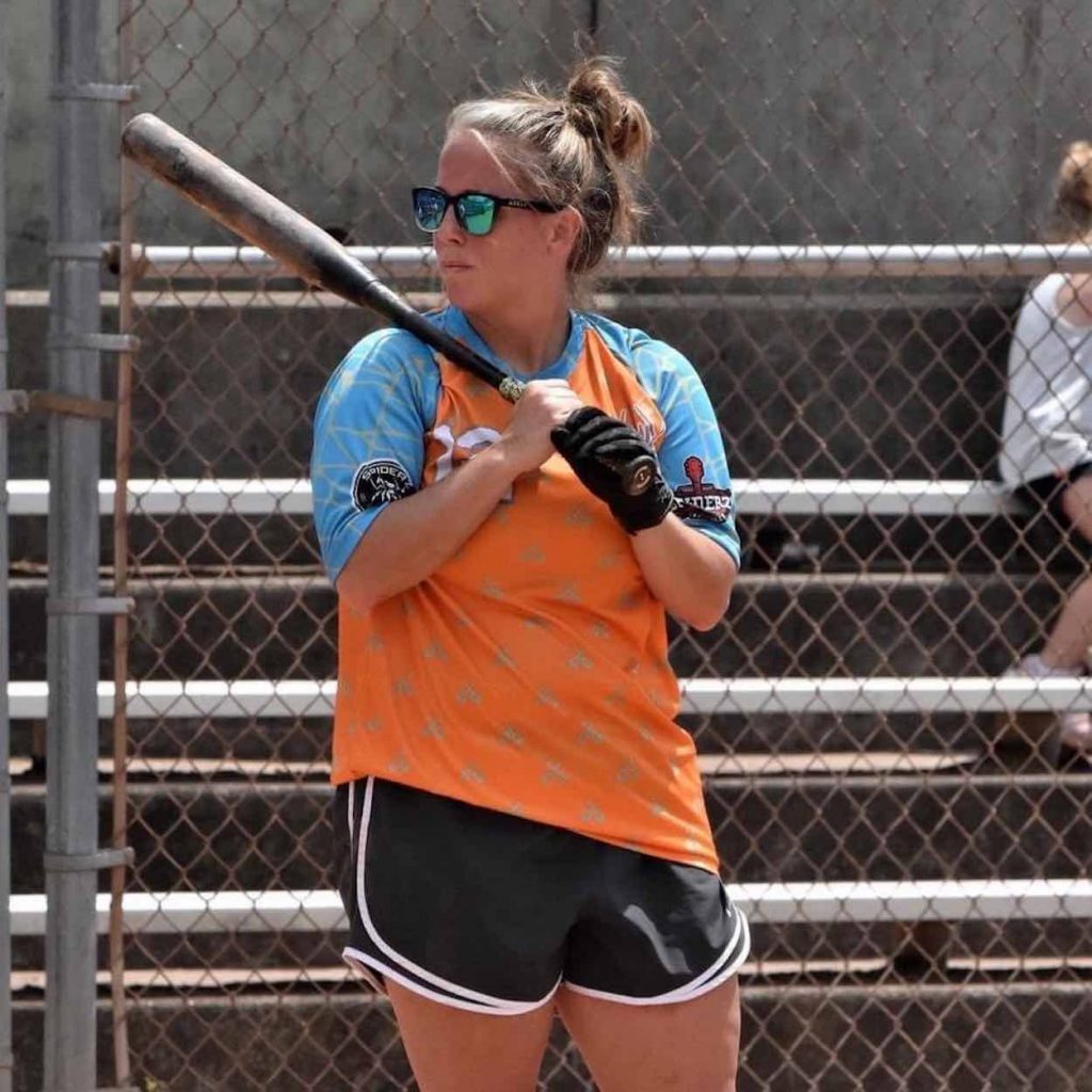 Jori is holding a bat and wearing an orange shirt as she plays softball.
