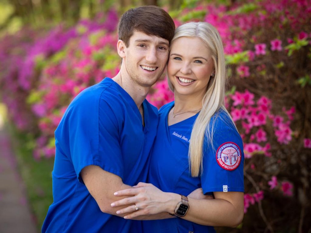 Heine and Hutto, both wearing blue scrubs, pose in front of a pink azalea bush. 