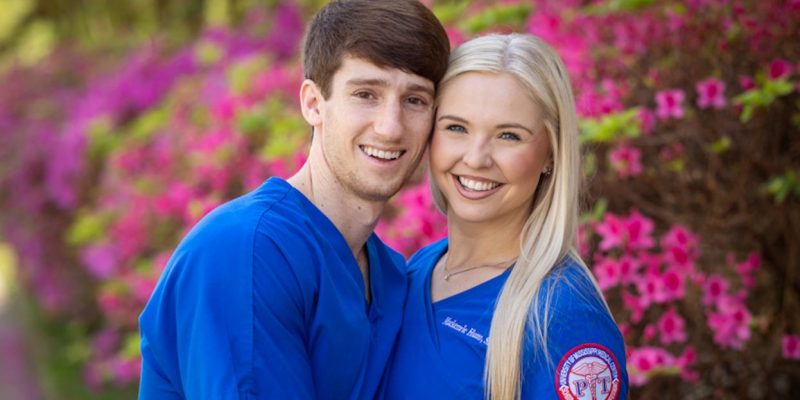Heine and Hutto pose in blue medical scrubs in front of a flowering tree.