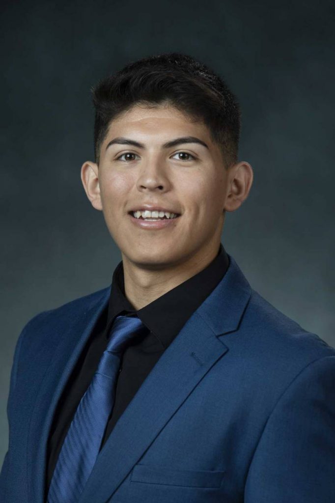 Miguel is in a blue blazer, blue tie and black shirt for his headshot photo. 