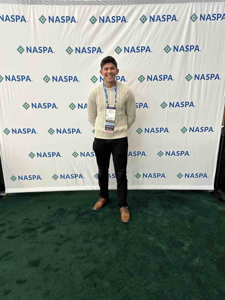 Miguel stands smiling in a white sweater with black pants in front of pop up back drop with the NASPA logo.