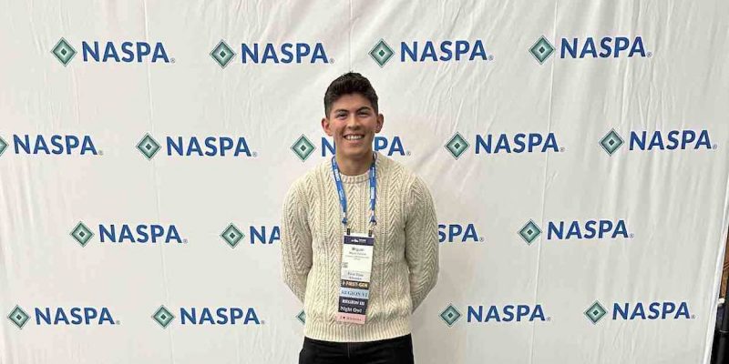 Miguel stands smiling in a white sweater with black pants in front of pop up back drop with the NASPA logo.