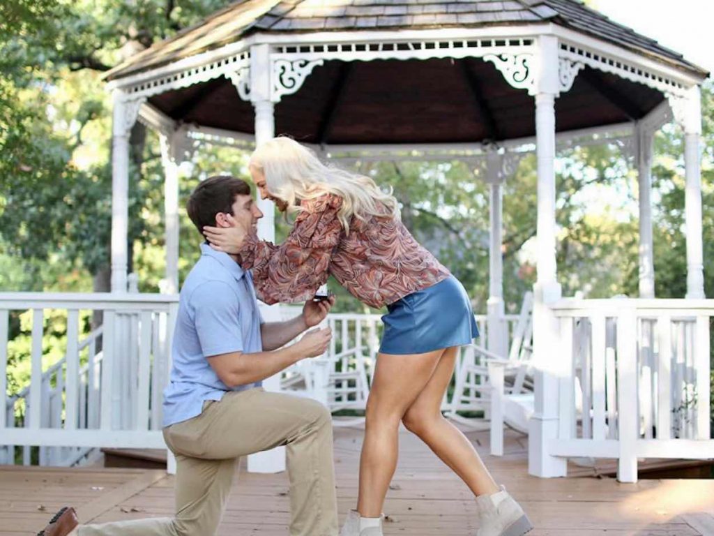 Heine is down on one knee proposing to Hutto in front of a gazebo. 