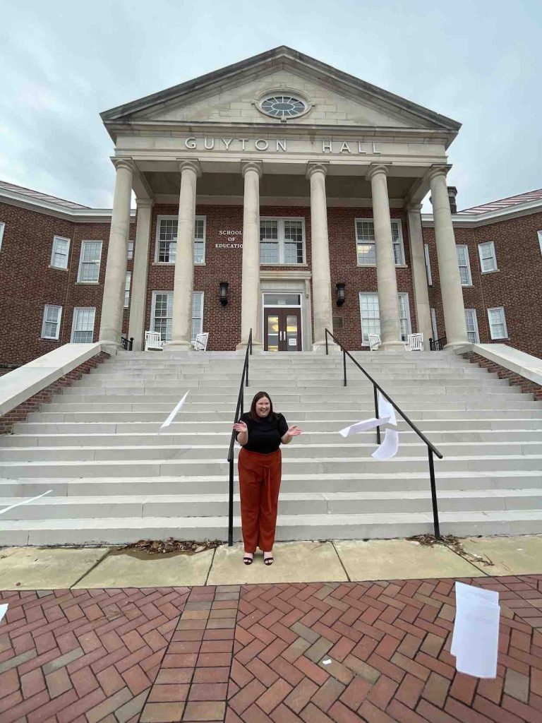 Raegan wears a black shirt and rust-colored pants as she throws the papers of her thesis in the air in front of the stairs of Guyton Hall. 