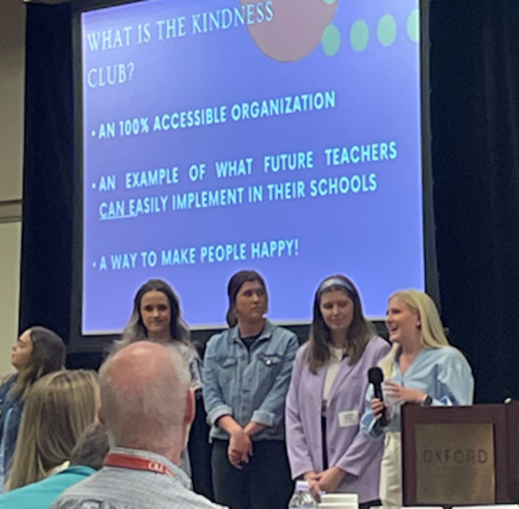 Abby stands to the left of a podium, holding a microphone in front of a screen with a presentation about the Kindness Club. Three other students are standing next to Abby as an audience watches.