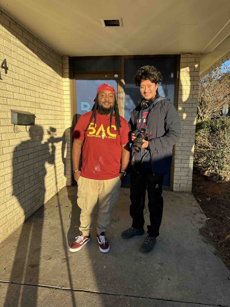 Sandip holds a camera in his hands as he stands next to a man in a red shirt and khaki pants outside of a white brick building. 