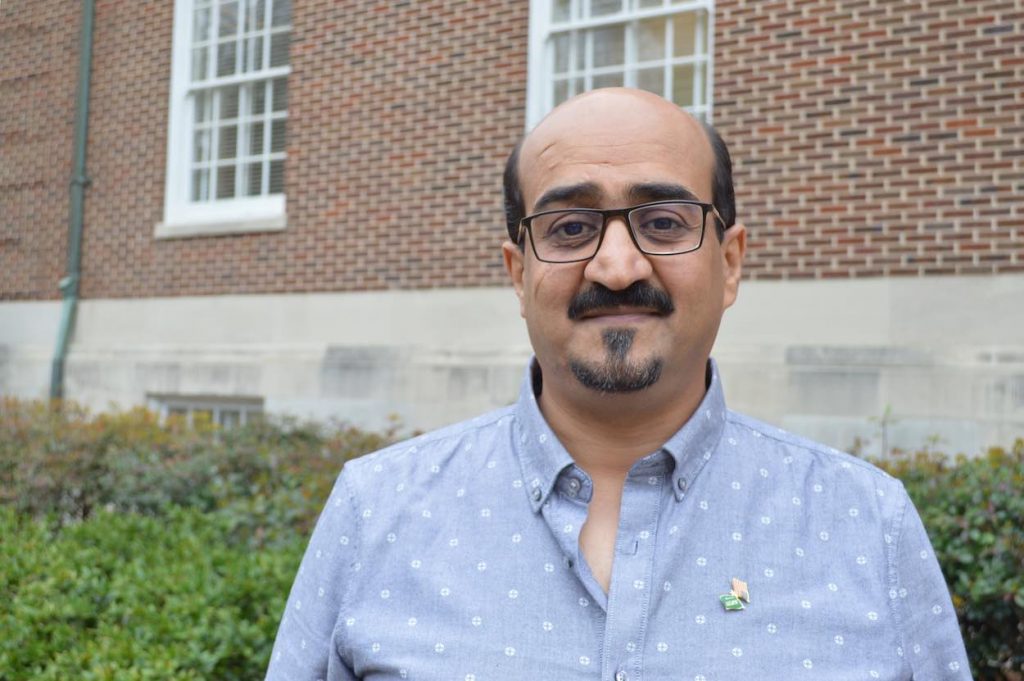 Abdullah stands in black glasses and a blue shirt in front of a brick building.