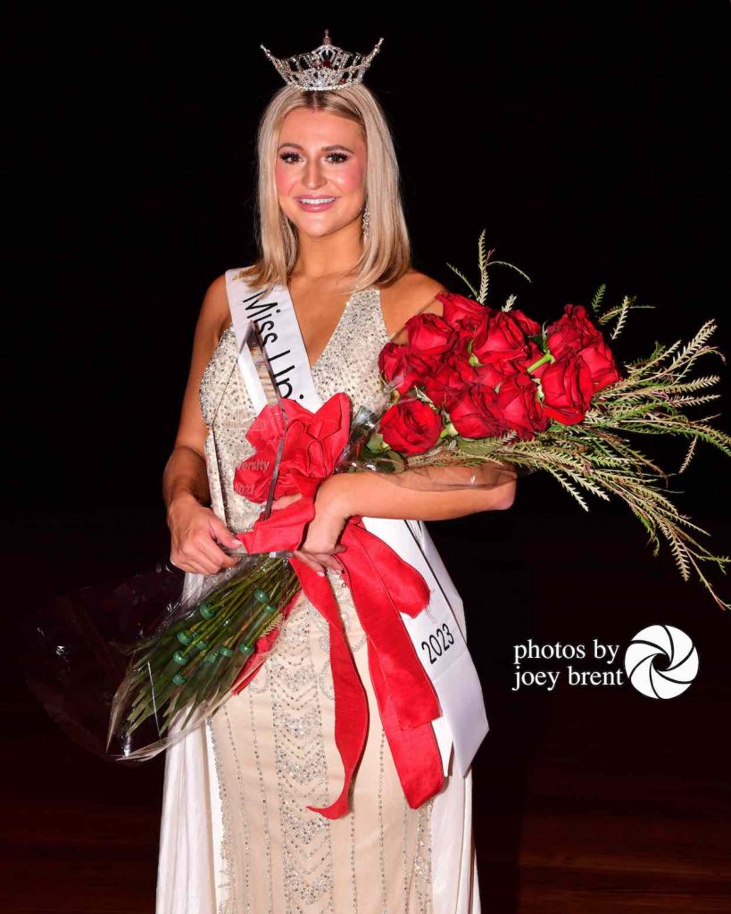 Hopper stands in a formal dress and crown with a Miss University sash across her front and red roses in her arms.