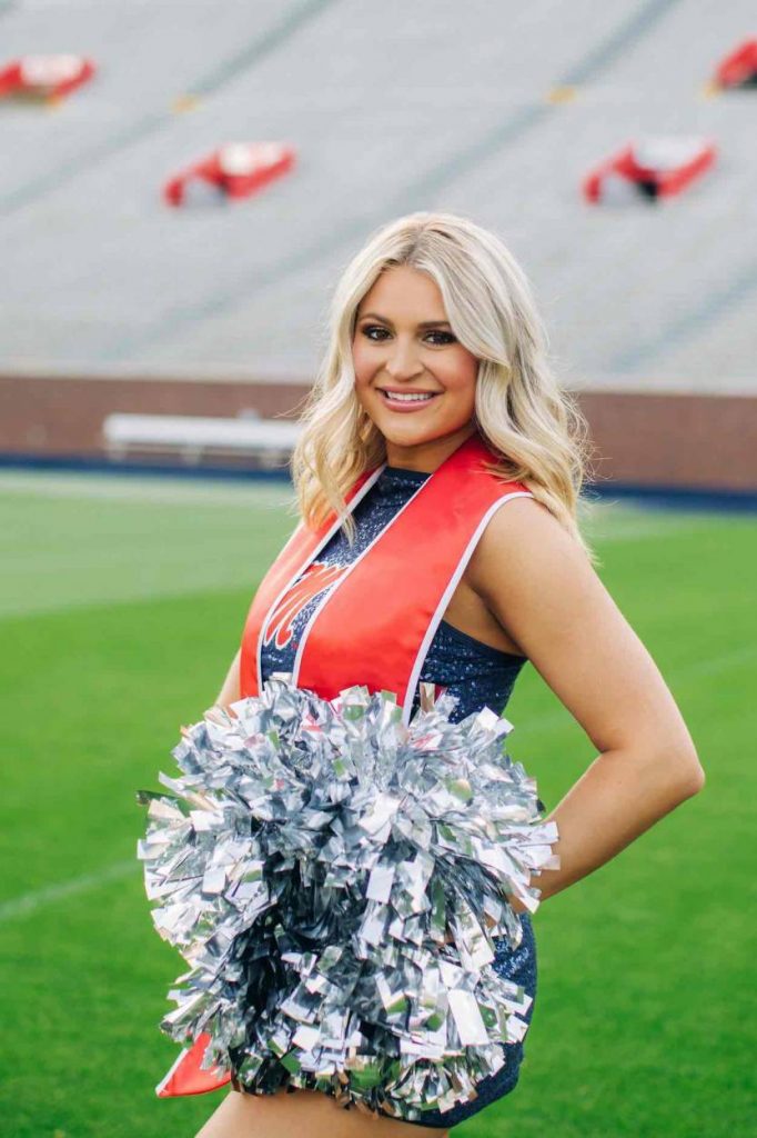 Hopper stands on a green football field in her blue Rebel dance uniform wearing a red graduation stole and holding silver pom poms.