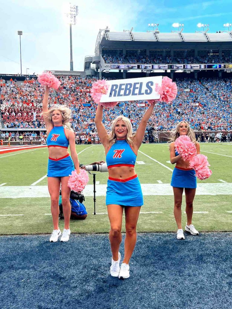 Three dance squad members stand on the football field in powder blue tops and skirts, holding pink pom poms.