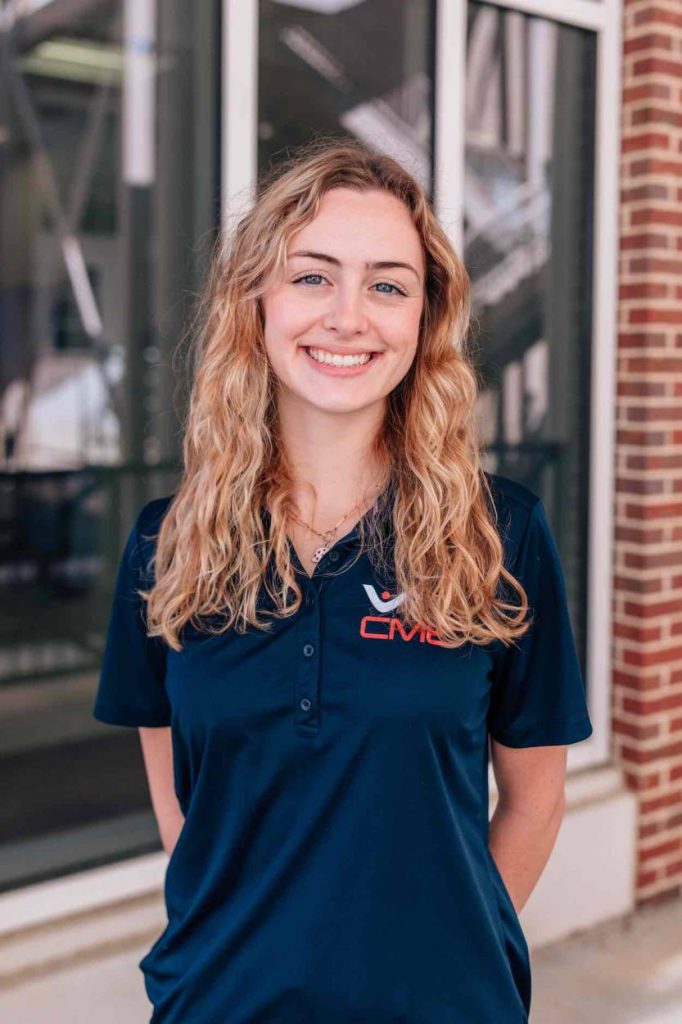 Millie stands in a blue collared shirt with the CME logo in front of a brick building.