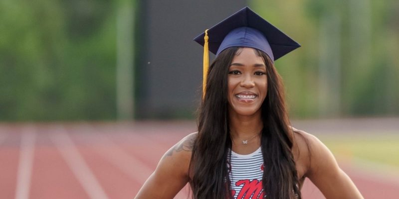 Orianna stands in her track uniform and graduation cap on a brick red running track.
