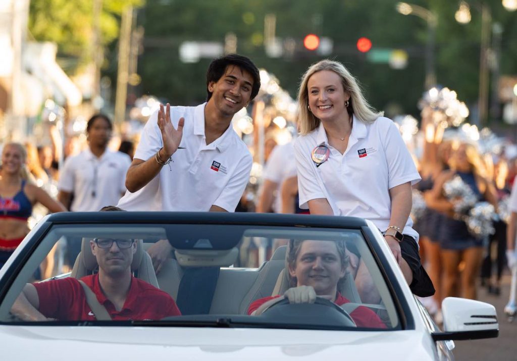 Emmaline wears a white collared shirt with an SAA logo in the backseat of a convertible with the top down for the Homecoming parade. 