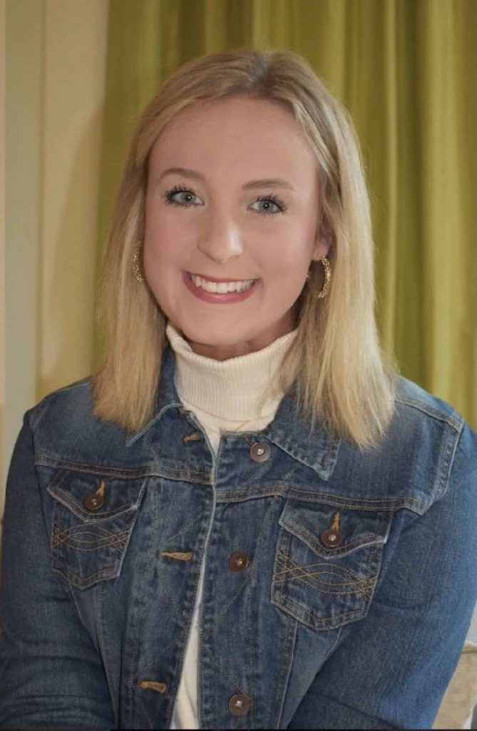 Emmaline poses in a cream turtle neck and jean jacket in front of yellow backdrop. 