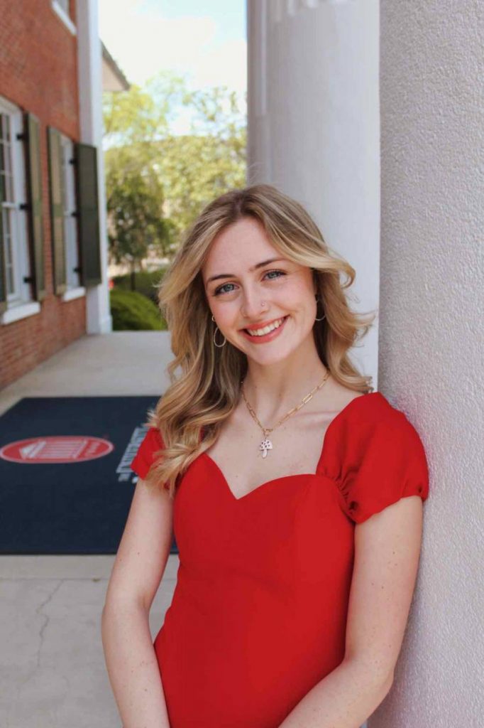 Millie in a red dress leans against a white column in front of the Lyceum.