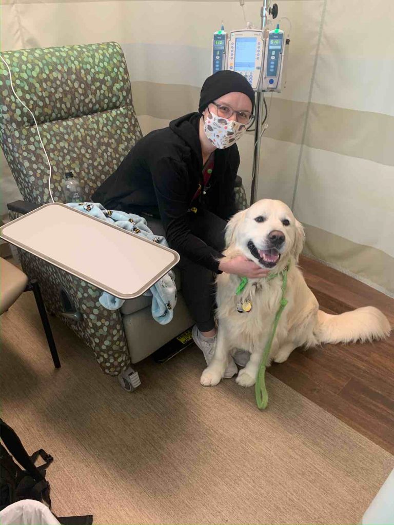 Heidi wears a knit cap and a mask as she sits in a treatment chair with a white dog near her. 