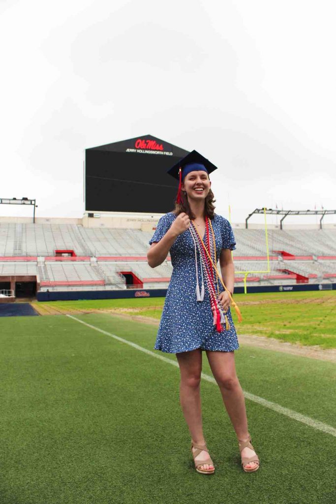 Heidi wears a blue dress and her graduation cap and cords as she poses on a green football field with stands and a jumbo tron behind her. 