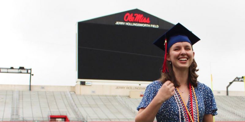 Heidi stands in a blue dress with her graduation cap on her head and her graduation cords around her neck. She is on green turf with the football stands and jumbotron in the background.