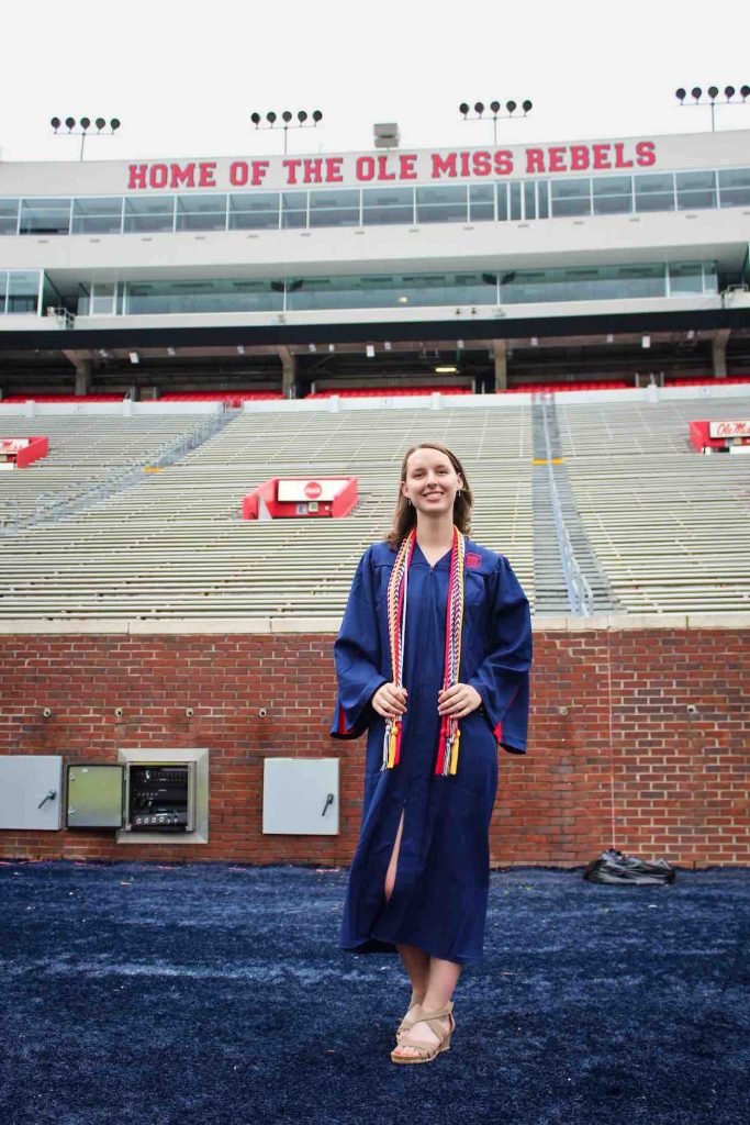 Heidi poses in a blue graduation gown with graduation cords around her next as she stands in front of football stands. 