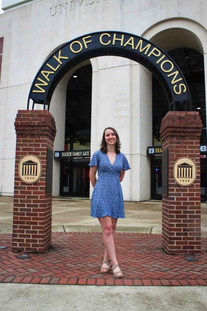 Heidi stands in a blue dress under an arch that says "Walk of Champions" in front of an entrance to the football stadium.