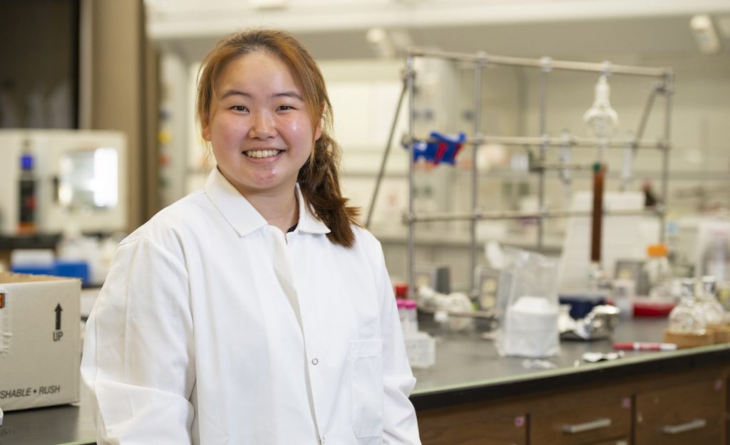 Suh is in a white lab coat with lab equipment behind her.