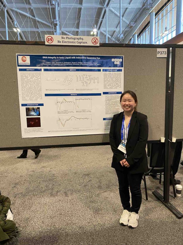 Suh stands in a black pants suit in front of a poster board presenting her research.