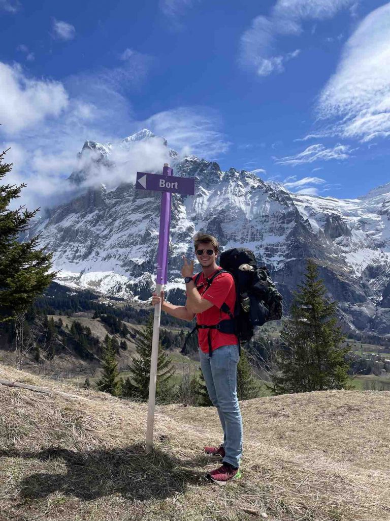 Matt is in a red tshirt and jeans in front a snow-capped mountain. 