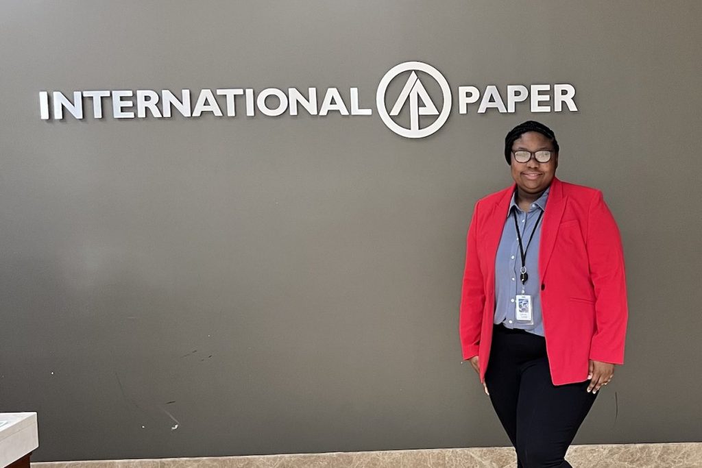 Casiah wears a red blazer and stands in front of a wall with the International Paper logo.