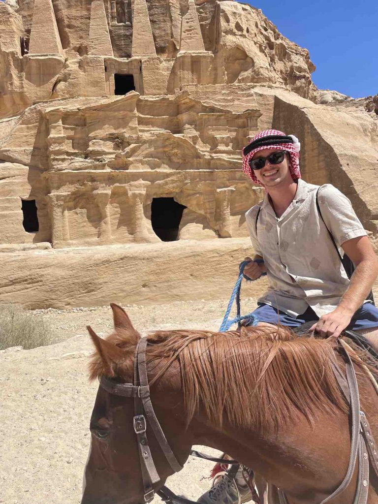 Matt wears a traditional red and white headdress worn by men from parts of the Middle East as he rides a horse in front of desert ruins. 