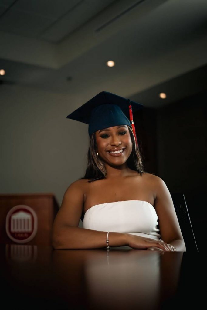Jilkiah sits at a table in a white dress and blue graduation cap with a red tassel.