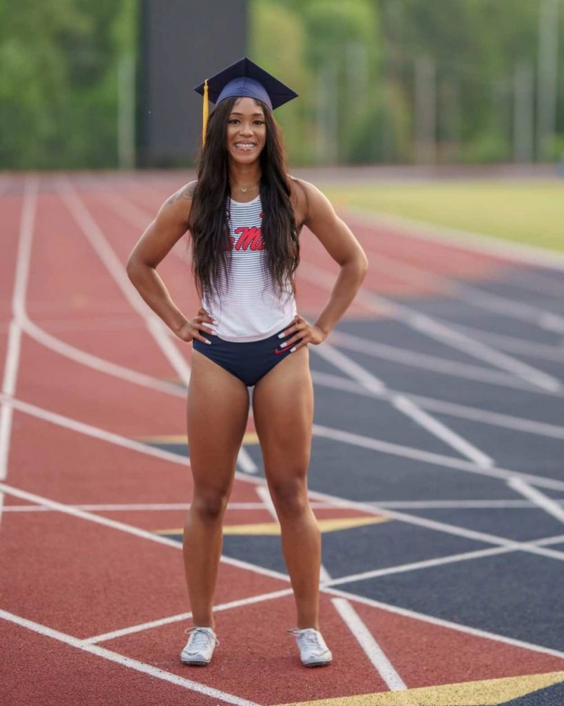 Orianna stands in her track uniform and graduation cap on a brick red running track. 