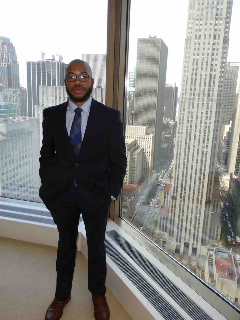 Jaylin stands in a dark suit in front of a large window with a wide view of New York City in the background.