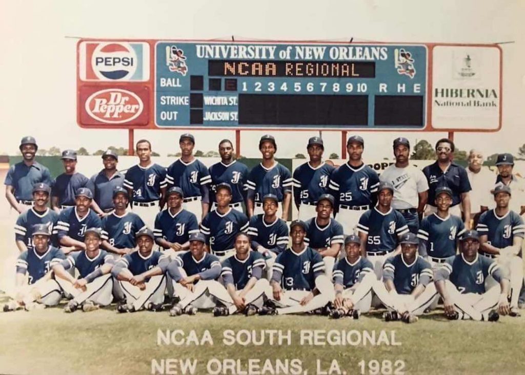 A team of baseball players pose in their uniforms in front of a score board.