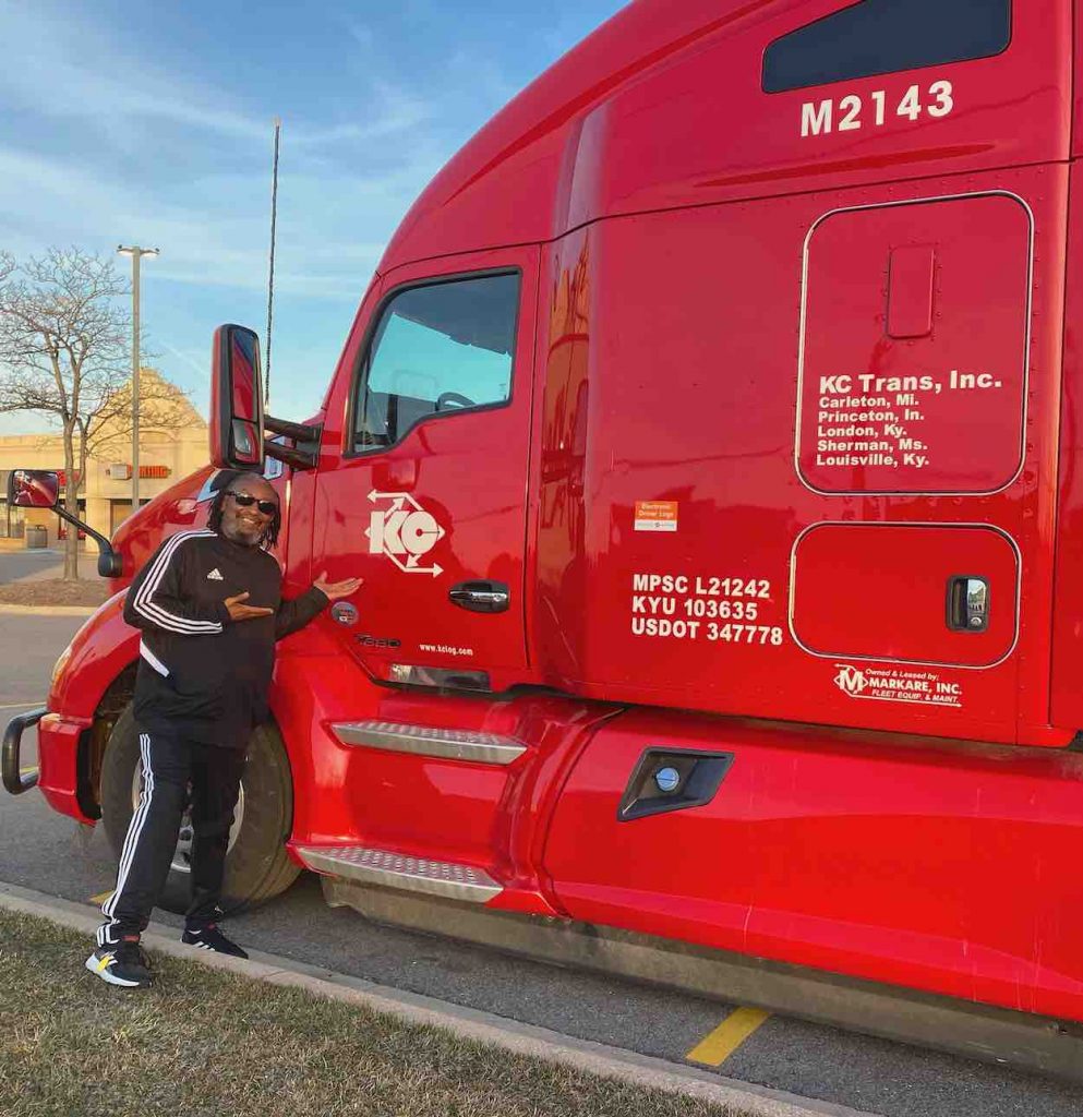 Leroy wears black pants and a windbreaker standing in front of a red 18-wheeler.