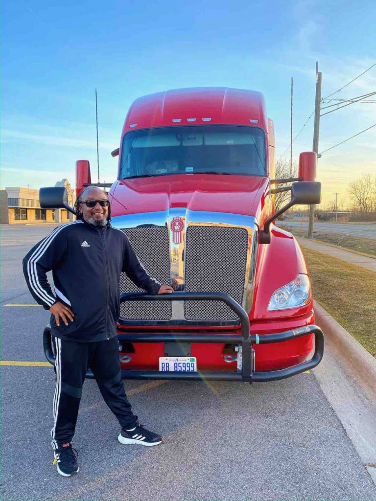 Leroy wears black pants and a windbreaker standing in front of a red 18-wheeler.