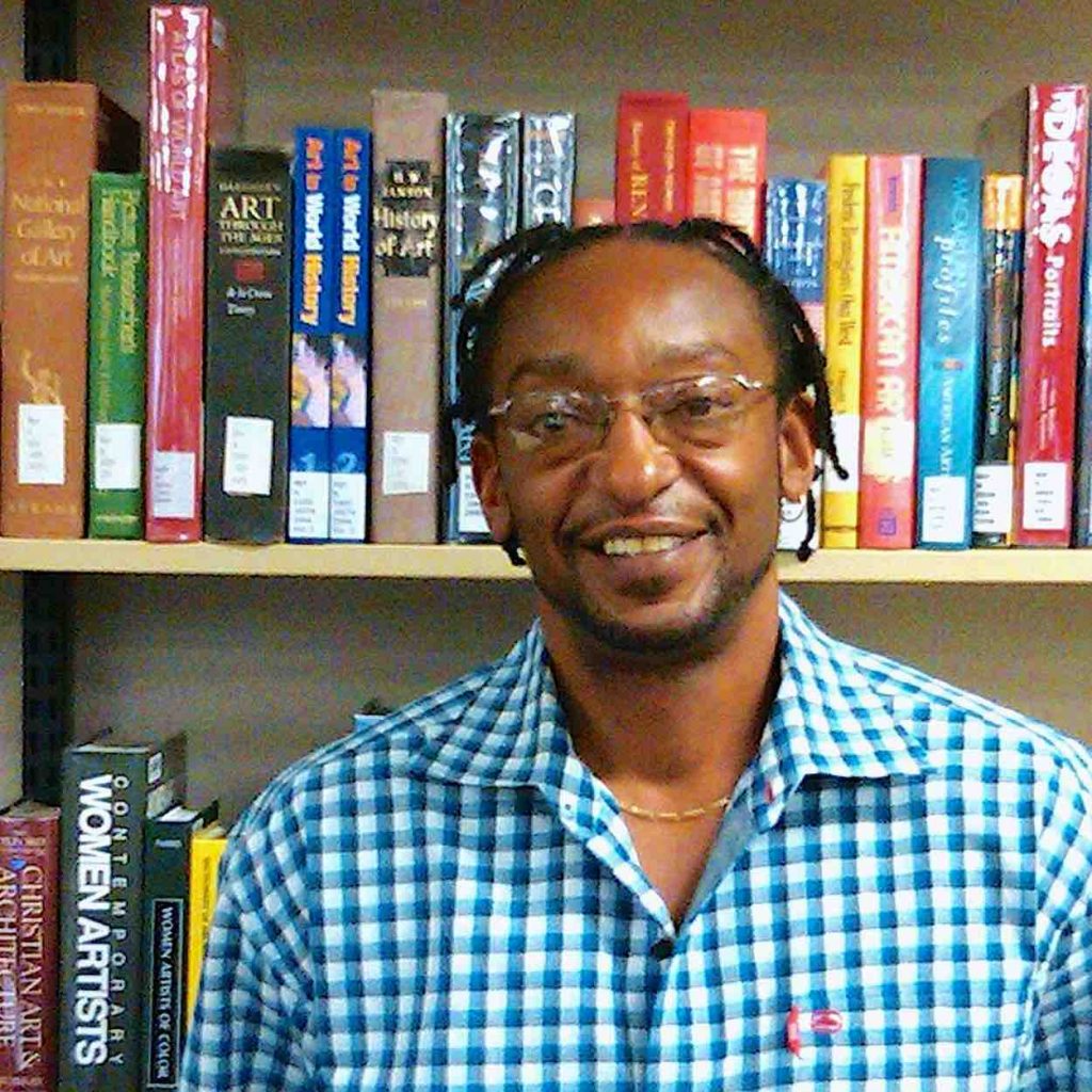 Leroy wears a blue checkered shirt in front of books on a shelf.