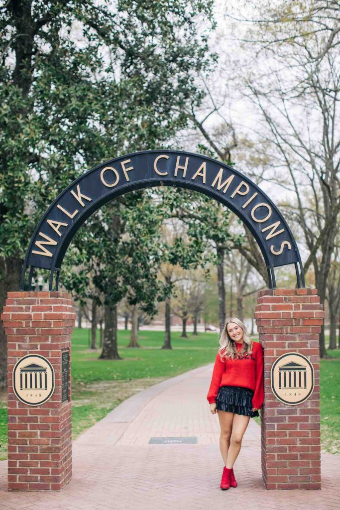 Mallory stands in a red sweater and black skirt under the Walk of Champions with the Grove in the background.