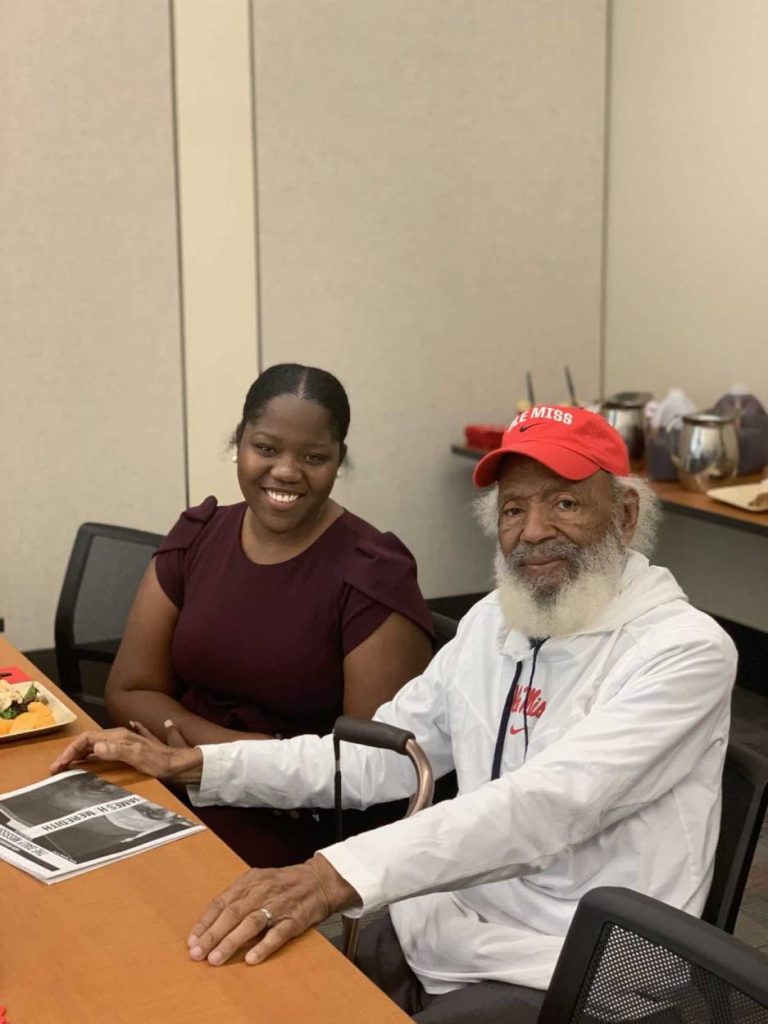 Moore wears a maroon dress as she sits next to James Meredith who is wearing a white pullover and a red Ole Miss hat.