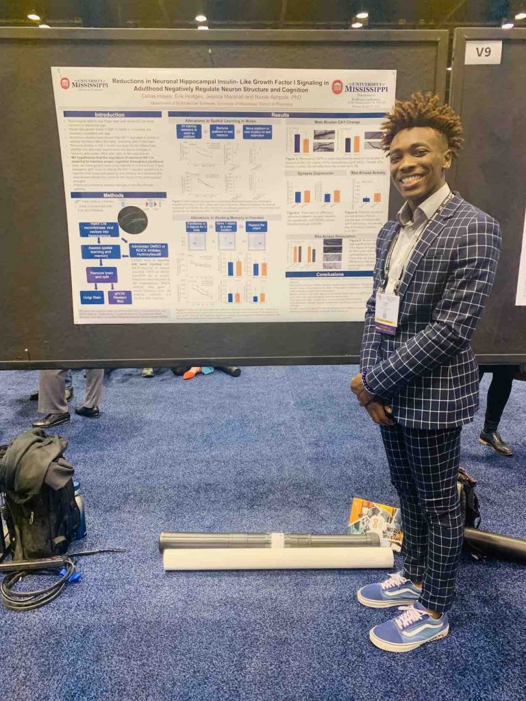 Hayes stands in blue pants and a blazer next to a research presentation board. 