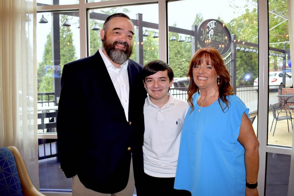 Rhonda in a blue shirt, her son in a white shirt, and her husband in a white shirt and dark blazer pose for a picture.