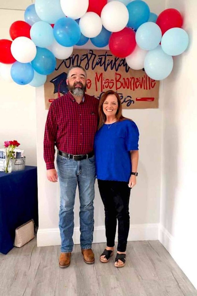 Brown in a blue shirt stands next to her husband in a maroon shirt in front of a homemade congratulations sign and red, white and blue ballons.